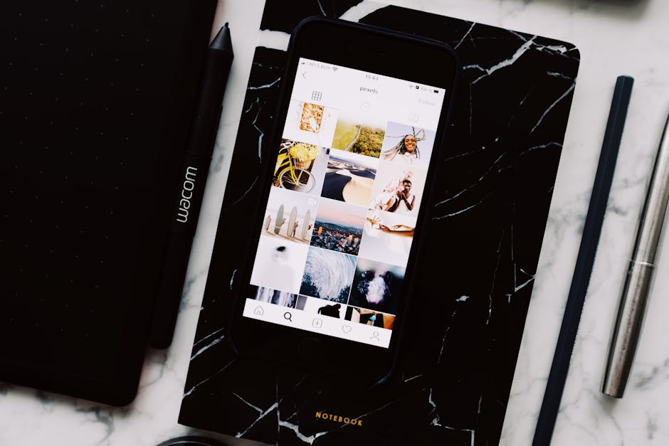 Top view of black notebook with cellphone above with pictures on screen and two pens aside located on marble table