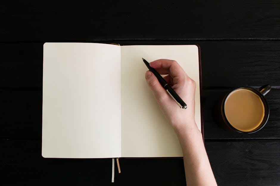Top view of a hand writing in a blank notebook with a coffee cup on a dark wooden table.