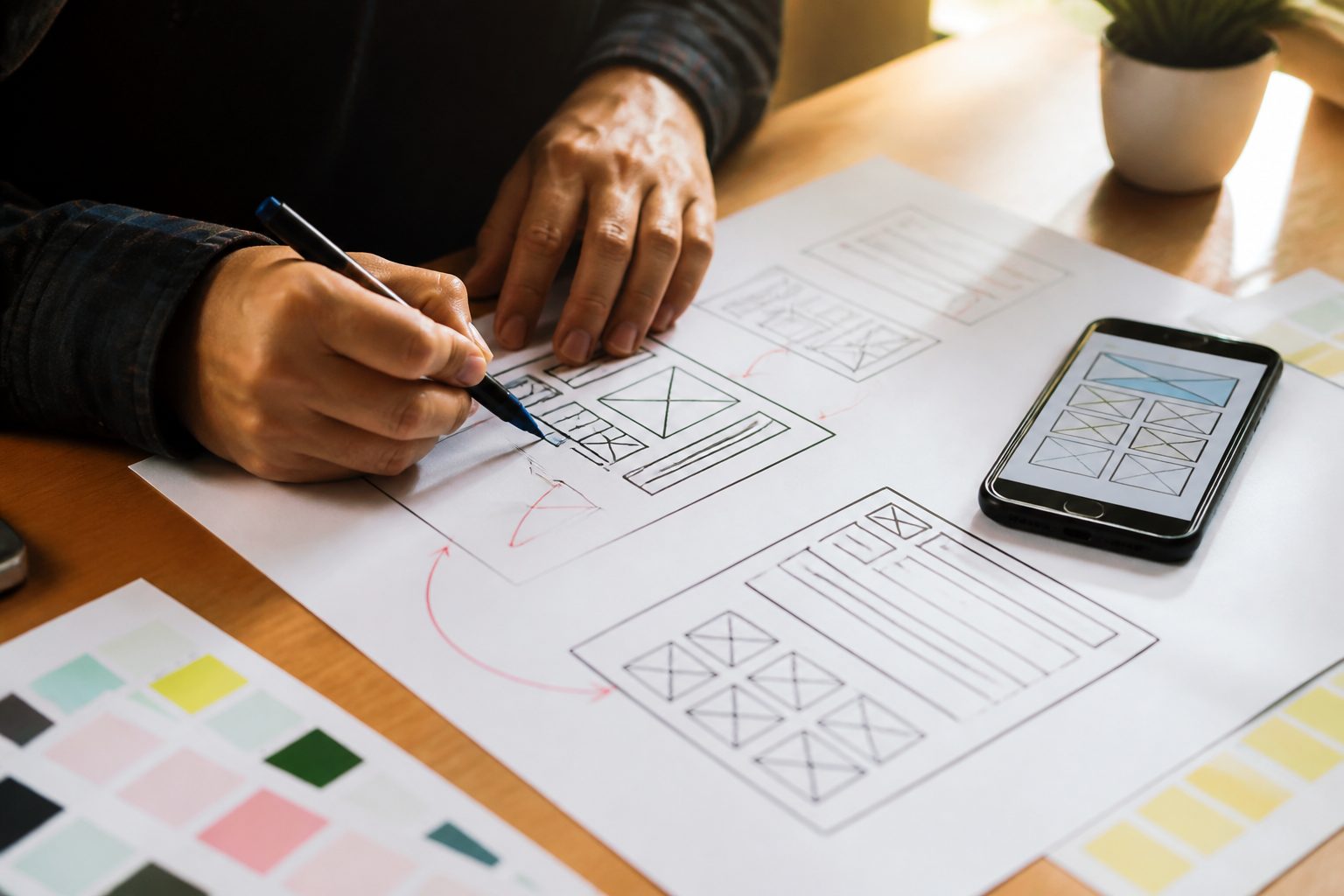 Close-up of hands sketching a website wireframe on paper at a desk, with a smartphone beside it showing a mobile layout mockup, warm natural light, focused and purposeful mood